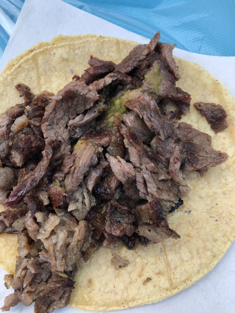 A close-up of a corn tortilla topped with grilled, sliced beef and a bit of green salsa, served on a white paper with a blue background—perfect for any travel blog exploring local flavors.
