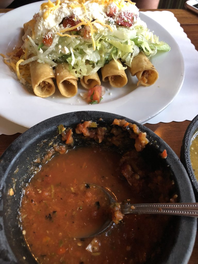 A plate of taquitos topped with lettuce, tomatoes, cheese, and sour cream, accompanied by a small bowl of salsa.