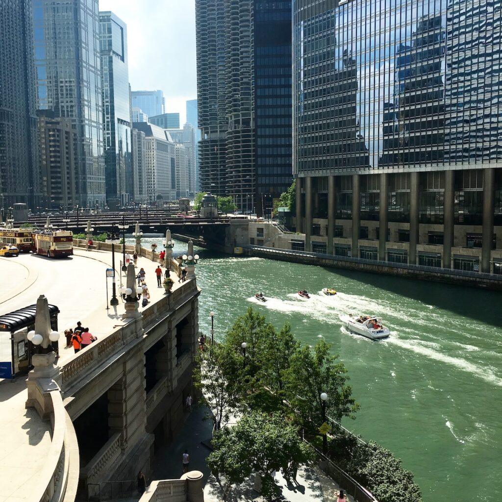A view of the Chicago River with several speedboats, surrounded by tall modern buildings; people walk along a riverside path and a bridge crosses over the water in the background—perfect for sharing on a travel blog like It's How I Travel.