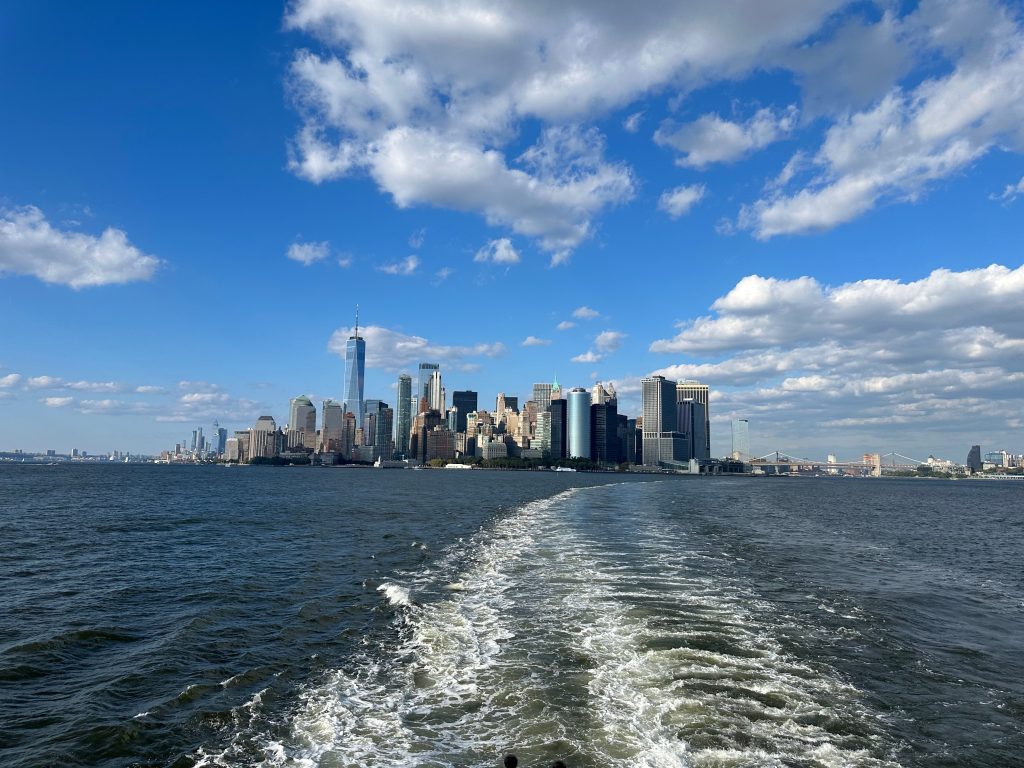 Experience the New York City skyline from the water, with a boat's wake leading you under scattered clouds—a moment captured for honest travel stories at It’s How I Travel.