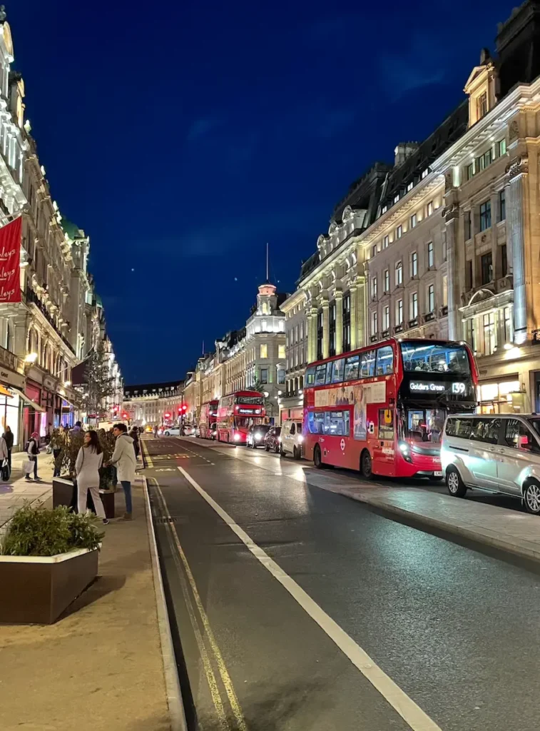 A busy city street at night in London, with red double-decker buses, cars, illuminated buildings, and pedestrians walking the sidewalk—capturing the vibrant energy of visiting London.
