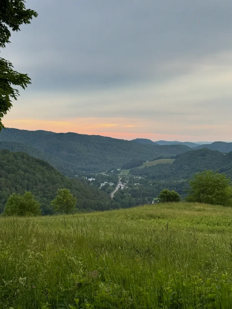 A green meadow overlooks a valley town surrounded by forested hills at sunset, with a cloudy sky tinged with orange at the horizon—capturing the serene charm often found in underrated states in the US.