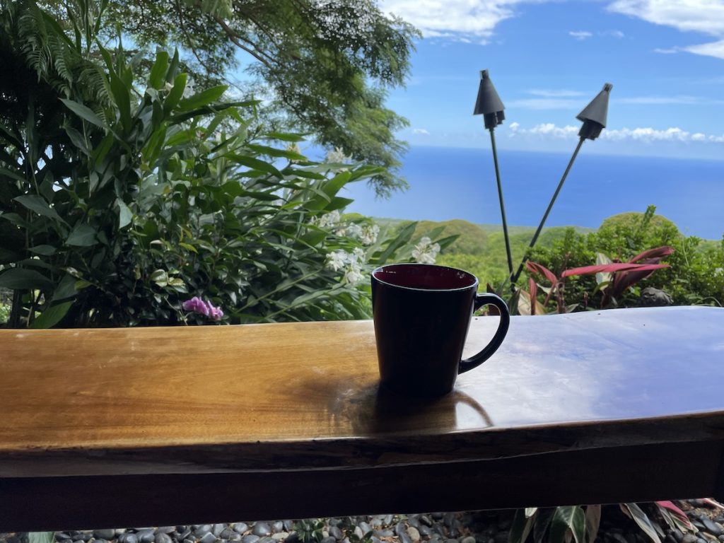 Coffee cup on a wooden table overlooking lush greenery and the Pacific Ocean in Kona.
