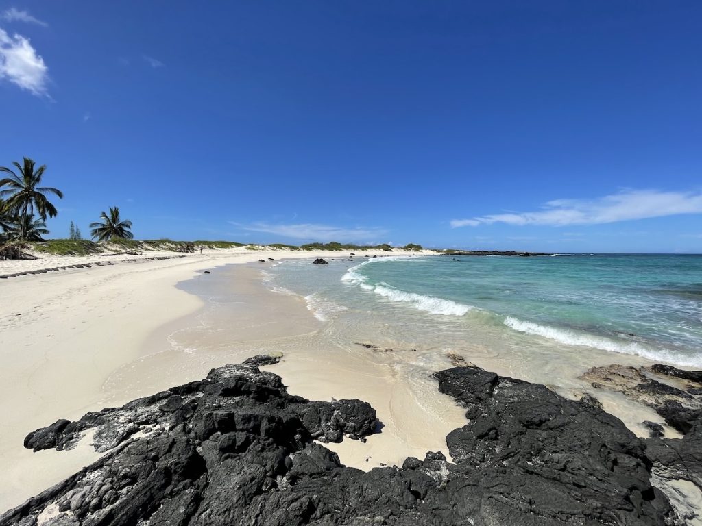 White sand and turquoise waves at Makalawena Beach with black lava rocks in the foreground.