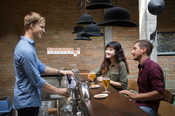 A bartender smiles while serving drinks to a woman and a man at a bar, with glasses of beer and small plates on the counter. The cozy, modern setting with brick walls is perfect for sharing travel stories—it's how I travel.