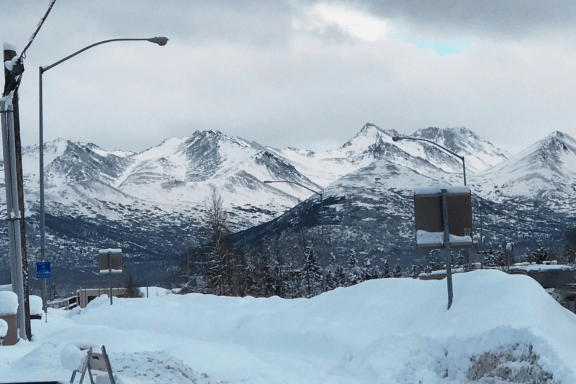 Snow-covered mountains under a cloudy sky, with a street in the foreground lined with snowbanks, street signs, and lamp posts capture the serene beauty of Alaska in January. Leafless trees stand between the road and the mountains.