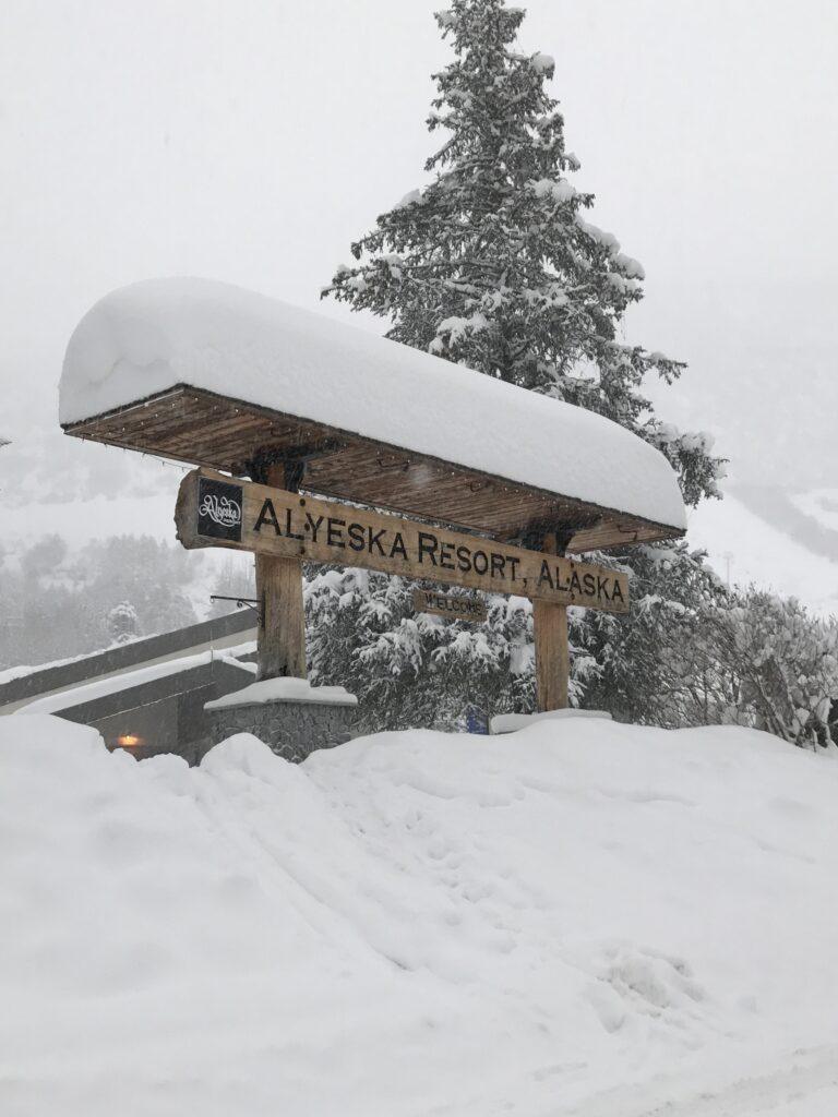 A wooden sign reading Alyeska Resort, Alaska stands under a snow-covered roof, surrounded by heavy snow and evergreen trees on a wintry Alaska in January day.