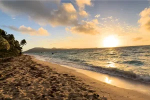 Golden sunset over a sandy Fiji beach with gentle waves, palm trees on the left, and distant mountains—perfect for a tranquil village homestay under a partly cloudy sky.