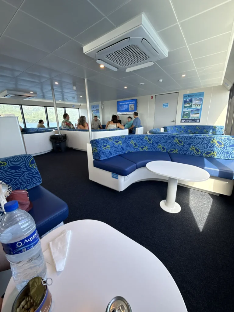 Interior of a ferry with blue patterned seats and white tables. Several people, perhaps sharing stories from a Fiji village homestay, are talking in the background. A water bottle and napkins sit on a table near large windows flooding in natural light.