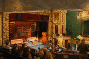 A crowded theater in Wilmington NC, known for its vibrant entertainment scene, features ornate golden columns and red curtains. From the balcony, the audience faces a stage set with benches and ladders, eagerly awaiting the performance.