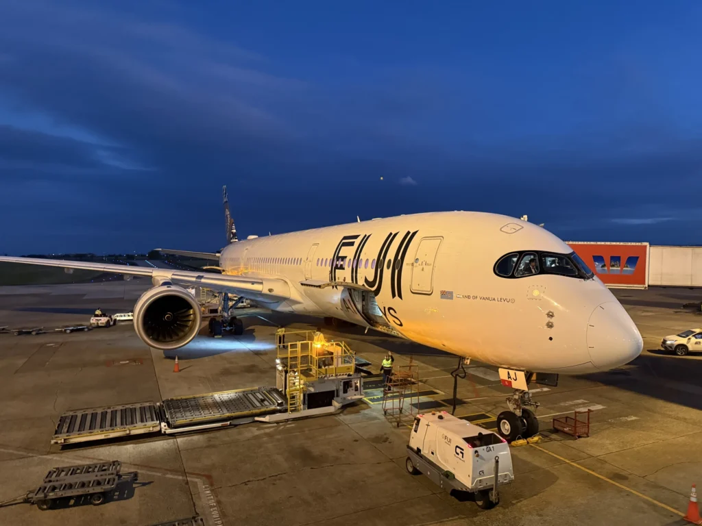 A Fiji Airways airplane is parked at NAN airport during sunrise, as ground crew and equipment ready the plane for departure beneath a darkening blue sky—offering travelers a gateway to explore Fiji’s vibrant villages or enjoy a welcoming homestay experience.