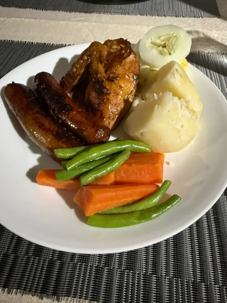 A white plate with roasted chicken wings, sausages, boiled potatoes, sliced cucumber, carrots, and green beans on a woven placemat—perfect for a cozy Fiji village homestay meal.