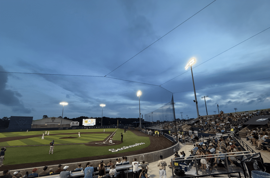 A baseball game at a stadium in Wilmington NC during dusk, with players on the field, spectators filling the stands, and stadium lights illuminating the scene under a cloudy blue sky—a perfect evening of entertainment in this vibrant beach town.