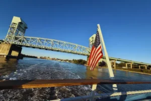 An American flag waves at the back of a boat on a river in Wilmington NC, with a large steel drawbridge and clear blue sky in the background. The water shows the boat’s wake leading toward the bridge and local finds along nearby neighborhoods.
