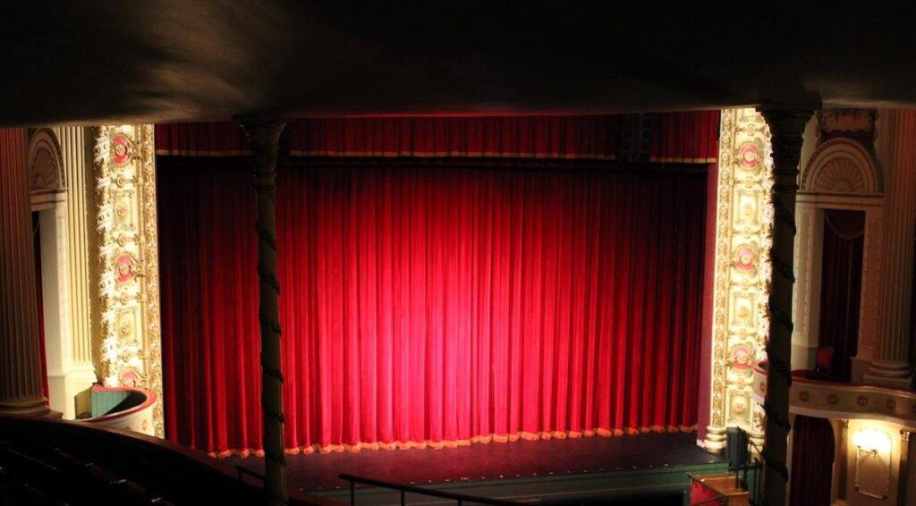 Thalian Hall, a theater stage with ornate columns on each side and a large, closed red curtain, viewed from the audience seating area under dim lighting—capturing the timeless charm of entertainment in Wilmington NC.