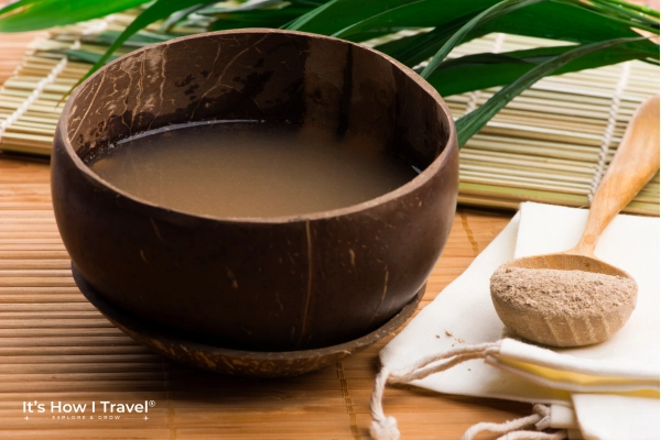 A wooden bowl filled with a cloudy brown liquid sits on a bamboo mat, next to a cloth with a wooden spoon containing light brown powder. Green tropical leaves evoke the relaxed charm of a Fiji village homestay in the background.