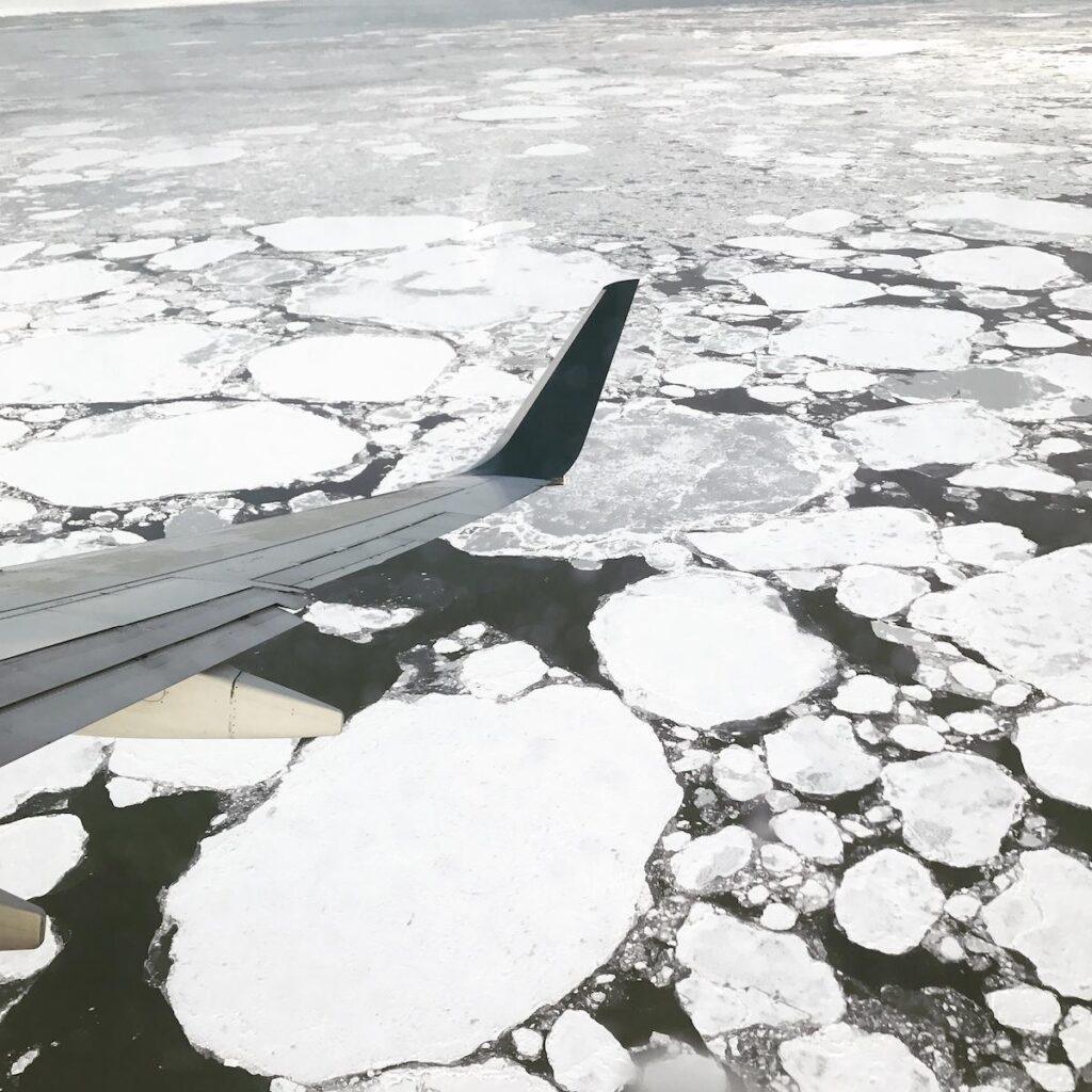 A view from an airplane window shows the plane’s wing and large circular sheets of ice floating on a dark body of water below, capturing the frozen beauty of Alaska in January.