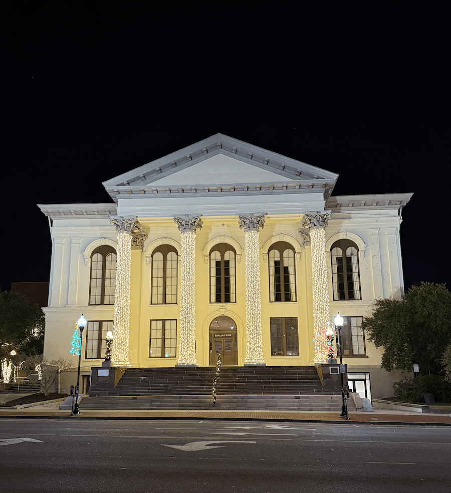 Thalian Hall. A large, neoclassical building with columns is illuminated by warm white string lights at night in Wilmington NC. Trees with lights and two colorful lit decorations stand near the entrance. The sky is dark, and the building is empty.