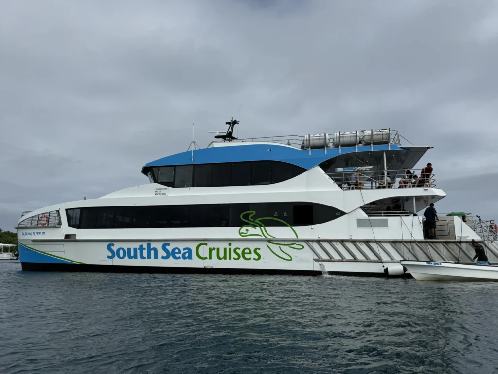 A large white and blue catamaran with South Sea Cruises written on the side is docked on calm water under a cloudy Fiji sky. Several people are visible on the upper and lower decks, ready to explore village life or stay at a local homestay.