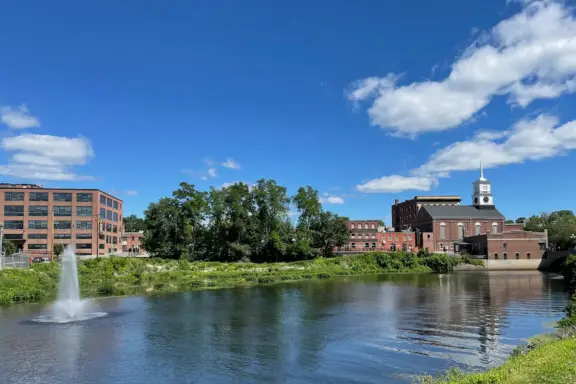 A calm river with a fountain, bordered by grassy banks, red-brick buildings, and a church with a white steeple under a bright blue sky with scattered clouds—quintessential scenery in New Hampshire, often regarded as the best state for tranquil beauty.