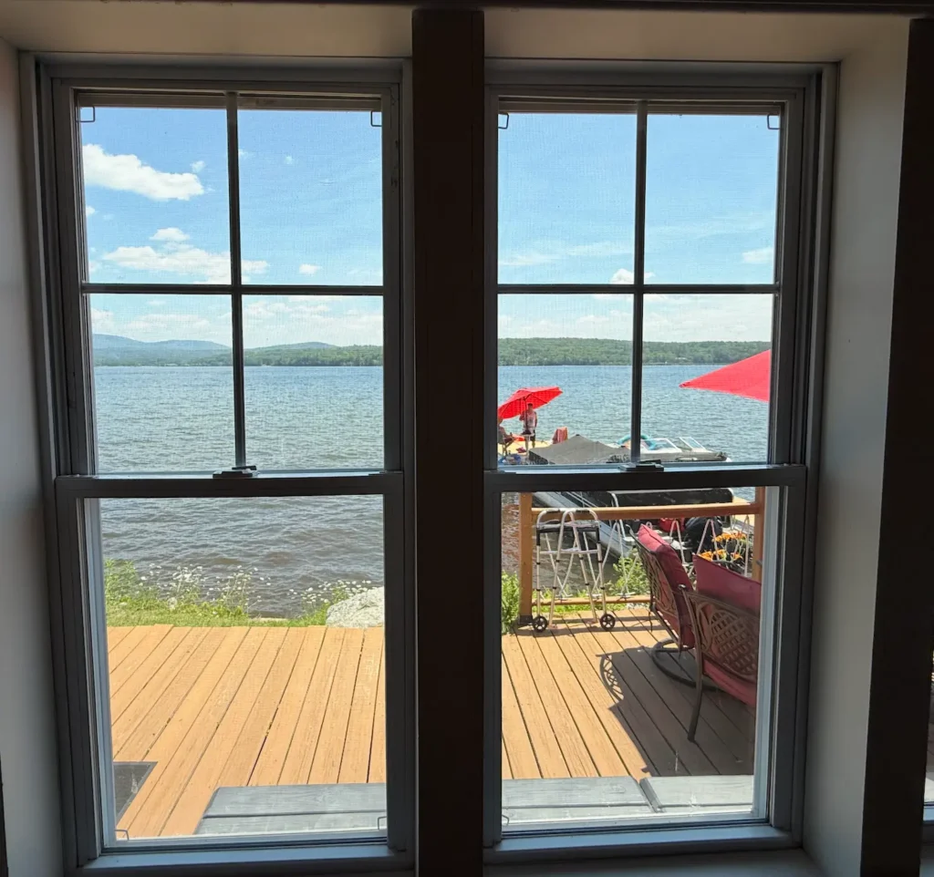View through a window showing a deck with red umbrellas and chairs, overlooking a lake with mountains and a blue sky in the background—capturing the serene beauty that makes New Hampshire one of the best states for nature lovers.