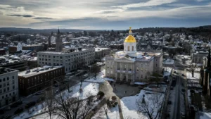 Aerial view of a grand building with a gold dome, surrounded by snow-covered grounds and city buildings under a cloudy sky in winter—inviting you to visit New Hampshire and experience its historic charm.