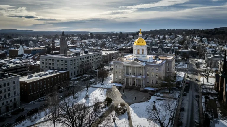 Aerial view of a grand building with a gold dome, surrounded by snow-covered grounds and city buildings under a cloudy sky in winter—inviting you to visit New Hampshire and experience its historic charm.