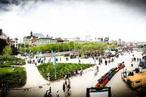 Busy urban park in Montreal with people walking, biking, and a small colorful train for children. Trees and greenery surround the area, with city buildings and a bridge visible in the background under a cloudy sky—perfect for inclusion in any Traveler's Guide to Canada's Most European City.