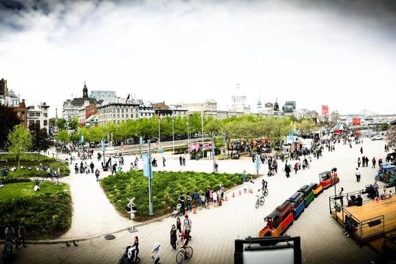 Busy urban park in Montreal with people walking, biking, and a small colorful train for children. Trees and greenery surround the area, with city buildings and a bridge visible in the background under a cloudy sky—perfect for inclusion in any Traveler's Guide to Canada's Most European City.