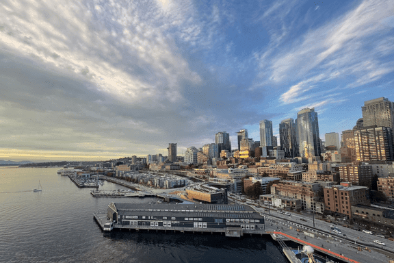 A city skyline with tall buildings next to a waterfront, a pier extending into the water, and a partly cloudy sky at sunset.