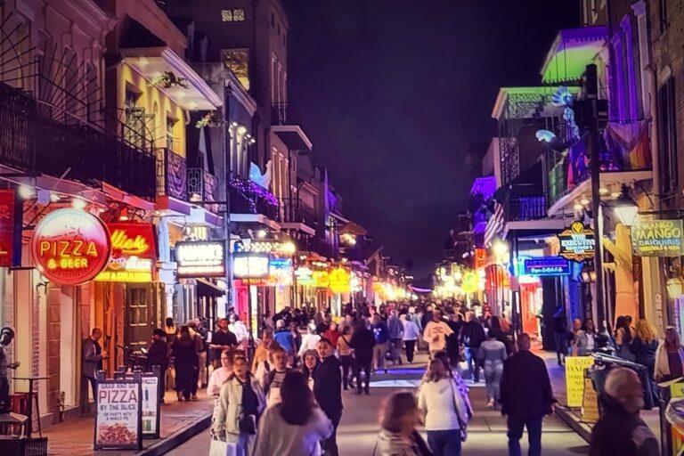 A busy Bourbon Street in New Orleans at night, crowded with pedestrians walking past illuminated shop signs, balconies, and restaurants.
