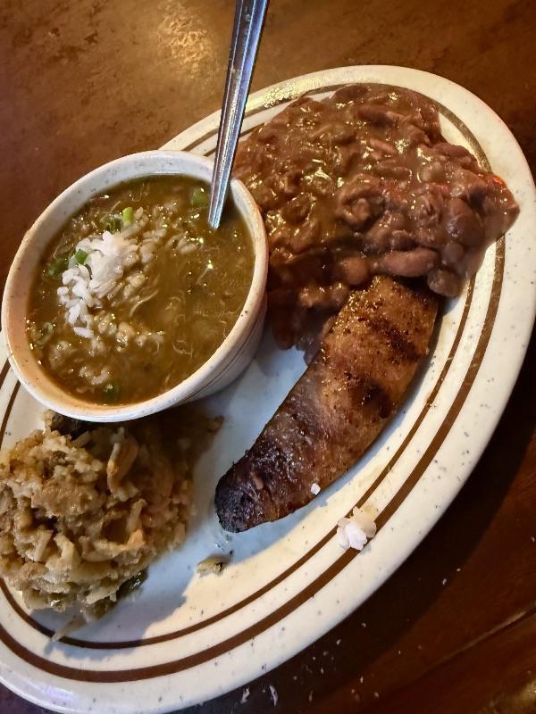 A plate of New Orleans food featuring a cup of gumbo, a scoop of brown beans, a serving of rice, and a piece of grilled sausage.