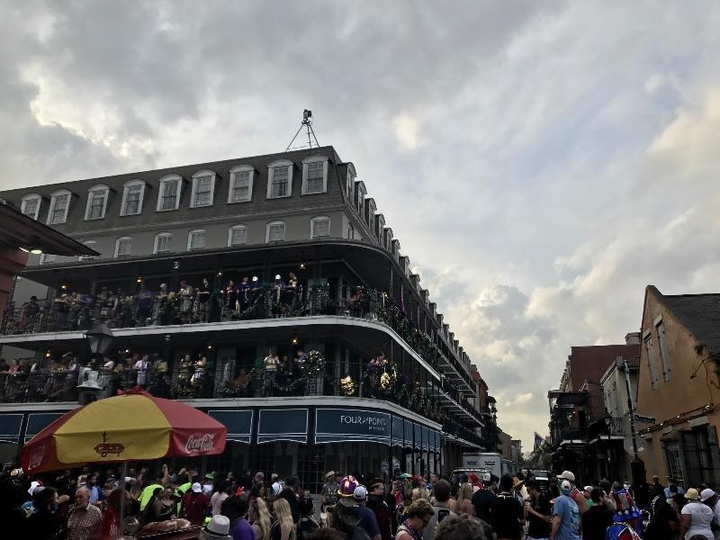 A crowd gathers on Bourbon Street beneath a multi-story building with balconies decorated for Mardi Gras in New Orleans.