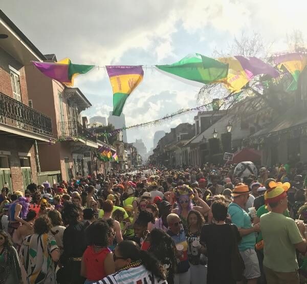A crowded Bourbon Street during Mardi Gras featuring colorful flags strung overhead, costumed revelers, and the traditional iron-balcony architecture of New Orleans.