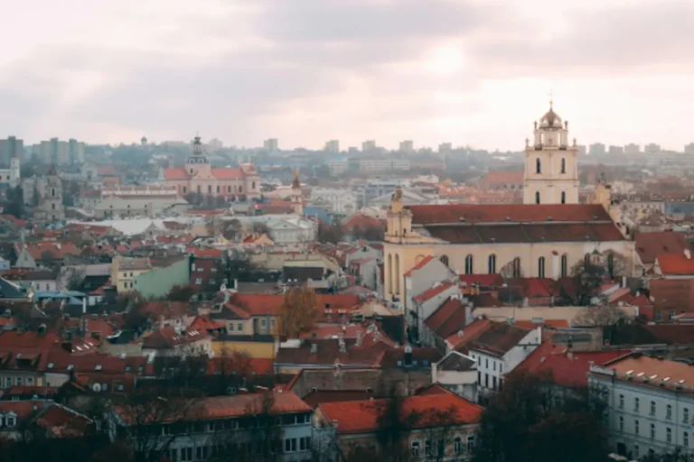 An aerial view of the red-tiled roofs and historic church towers of Vilnius, Lithuania, under a cloudy sky, showcasing the city’s unique charm for travel.