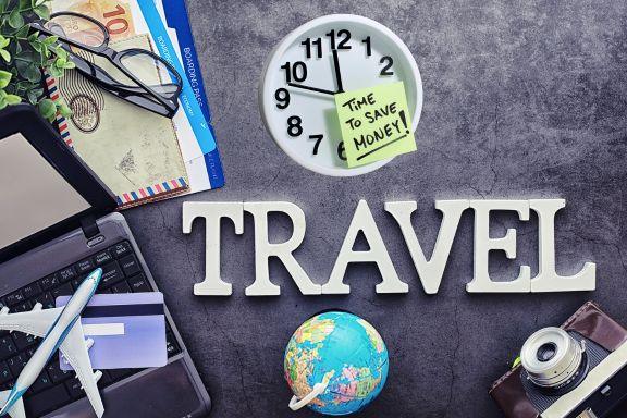 A workspace featuring a laptop, a model airplane, a credit card, a globe, a camera, travel documents, and a clock with a sticky note reading Time to save money on flights! above the word TRAVEL spelled out in large letters.