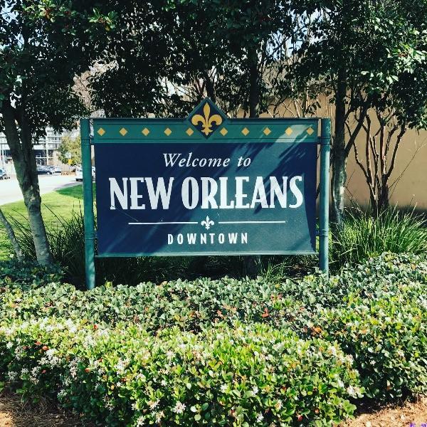 A Welcome to New Orleans Downtown sign with a gold fleur-de-lis emblem, set in a garden with trees and flowering shrubs often seen by those following a New Orleans travel guide.